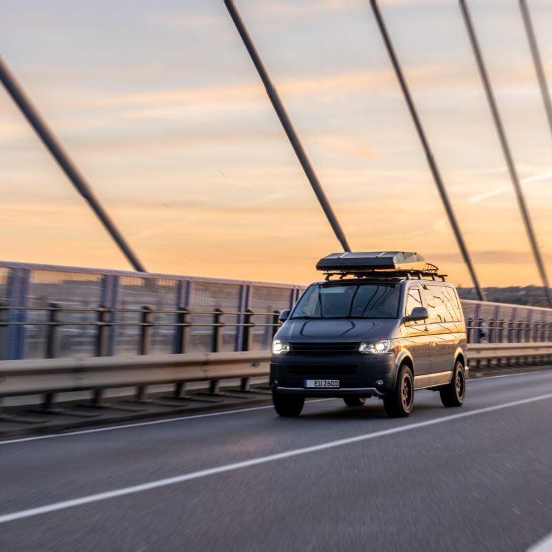 A dark modern minivan with roof box moves along the bridge at sunset light. Scene of freedom, travel and adventure with warm golden tones and urban landscape. SSUCv3H4sIAAAAAAAAA41Py46DMAz8F59BgqalwK9UPeThUqtAqsRstUL59zqUaq+bgzXjiT3jFchBD+h0Y92hKo26VWVdS2k748qDqlV1Ojat6QwU4DQj9LU6Vs25brtWnc6qkleA0ZEs9CvQOC6Rg2byM/SiBJwdhg2iI/aB9CgsFRBZ8xIxypgwK7sHUTf+3XdZc18CinlcTAap+GfvWoAecLa/eX/KQUbUm91FpMeLMUy7t+d7jpjhM5CledgzySl++uDZc57dHNjbB7l9+HPGnaQbshcsz9Frh26XFzMR85cG/CF8/f1OKb0BwPS5aYQBAAA=